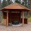 Wooden Wall to Gazebo Shelter՝ բացօթյա փայտե սրճարան այգու մեջ, շրջապատված ծառերով։